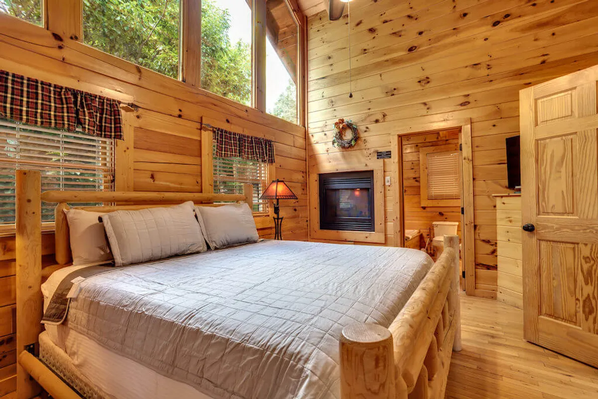 A cozy king bedroom featuring a log bed, a stone fireplace, large windows letting in natural sunlight, and a table lamp. The room exudes a warm, serene atmosphere perfect for relaxation and restful sleep.