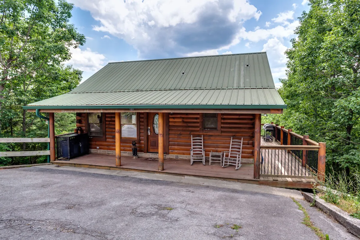 This image shows the exterior of a charming log cabin with a green metal roof, a wooden porch lined with rocking chairs, and lush trees surrounding the property. The inviting porch and natural setting capture the spirit of mountain living and the promise of outdoor adventure.