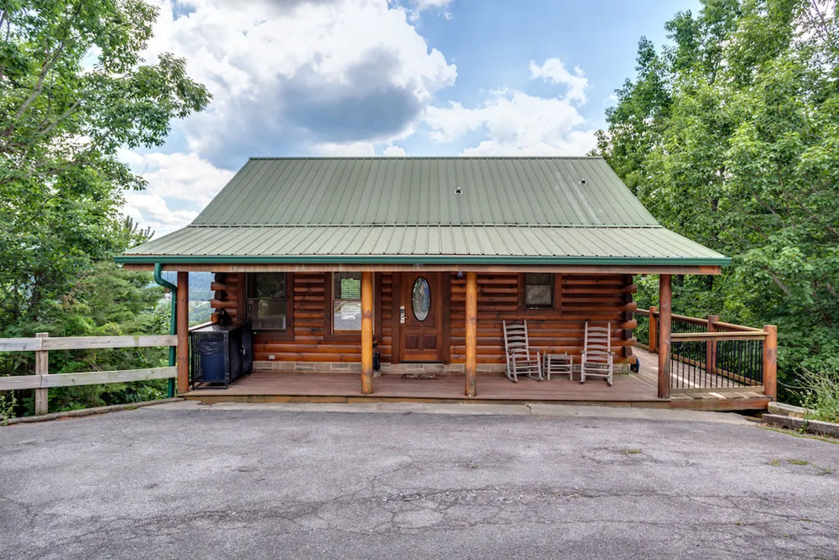 A classic log cabin with a green metal roof sits surrounded by lush trees. Its spacious deck features several rocking chairs, perfect for relaxing after a day of adventure. The inviting porch hints at family gatherings and peaceful evenings, while the expansive yard offers room to play and host outdoor activities.