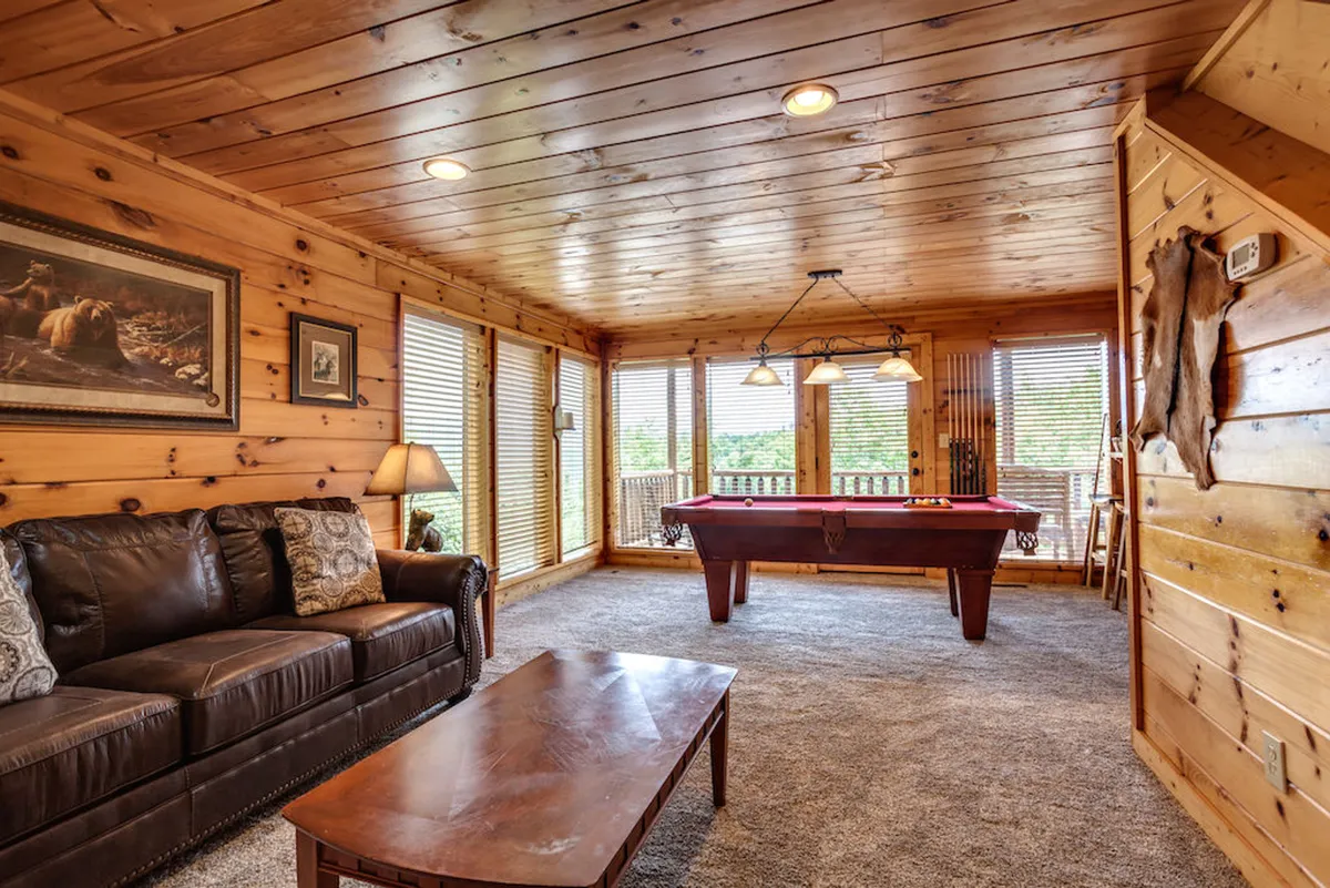 A casual and relaxed interior with a pool table in the foreground, a leather sofa, a wooden coffee table, and windows with blinds. The setup encourages friendly competition as well as restful moments between games.