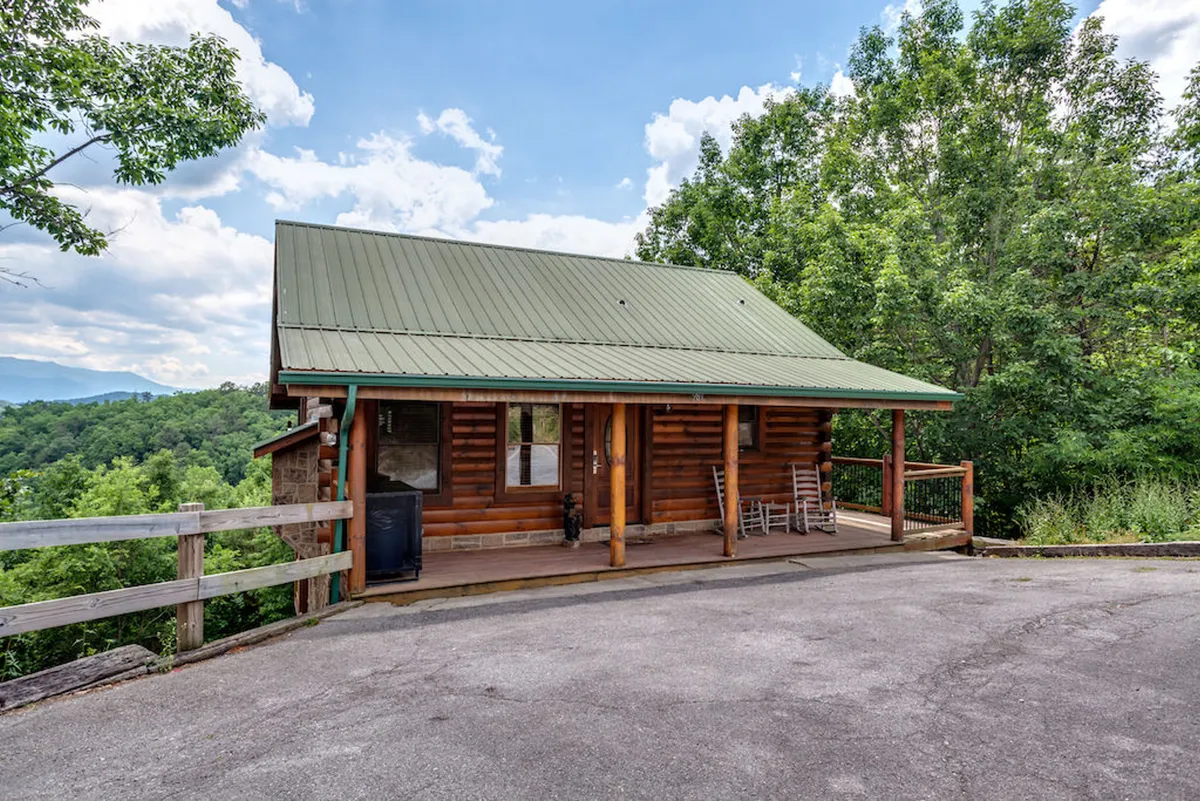 A charming log cabin with a green metal roof sits surrounded by lush greenery. Its welcoming wooden porch is furnished with classic rocking chairs, inviting guests to relax and enjoy nature. The tranquil setting hints at privacy and a strong connection to the natural world, echoing the sense of seclusion and romance highlighted in the section.