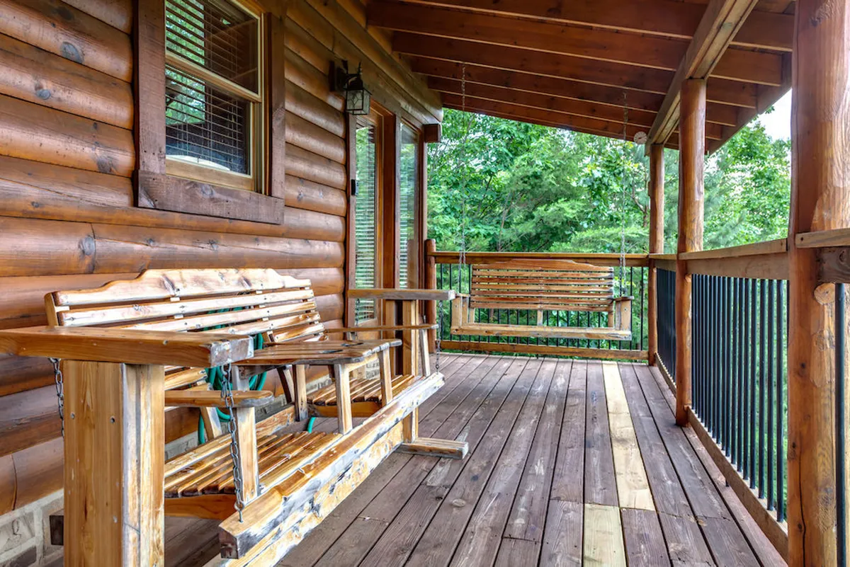 A cozy outdoor seating area features wooden benches and a swing set against the cabin’s log wall. Lush greenery spills into the scene, creating a private nook ideal for intimate moments, reading, or quiet reflection surrounded by nature.