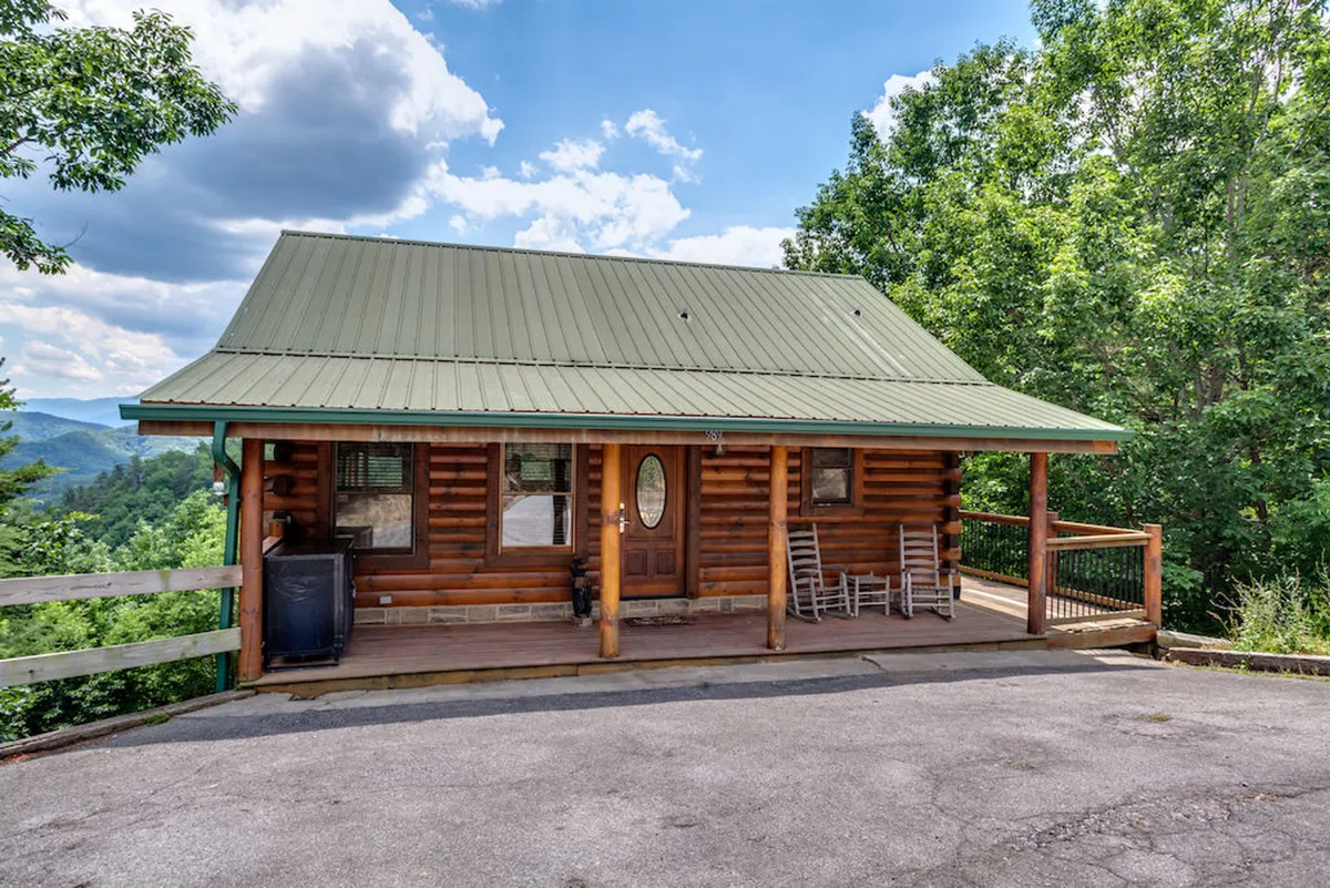 A welcoming log cabin with a green metal roof nestled among lush trees. The spacious front porch invites groups to relax or gather, providing an ideal setting for both morning coffee and sunset chats. The cabin exudes a cozy, rustic mountain charm, immediately setting the scene for a group retreat.