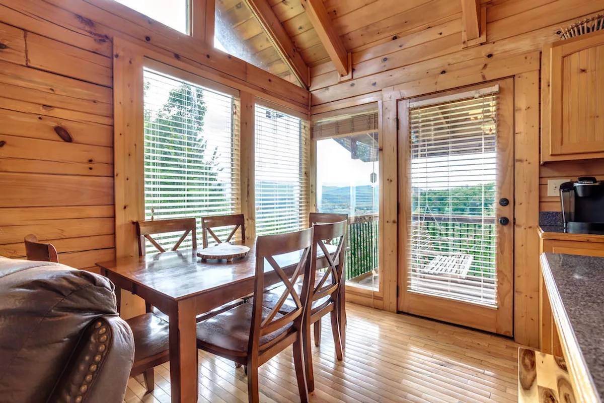 A wooden dining table bathed in natural light from large windows, with outdoor mountain views visible in the background. The setting is intimate, perfect for family meals, morning coffee, or evening board games.