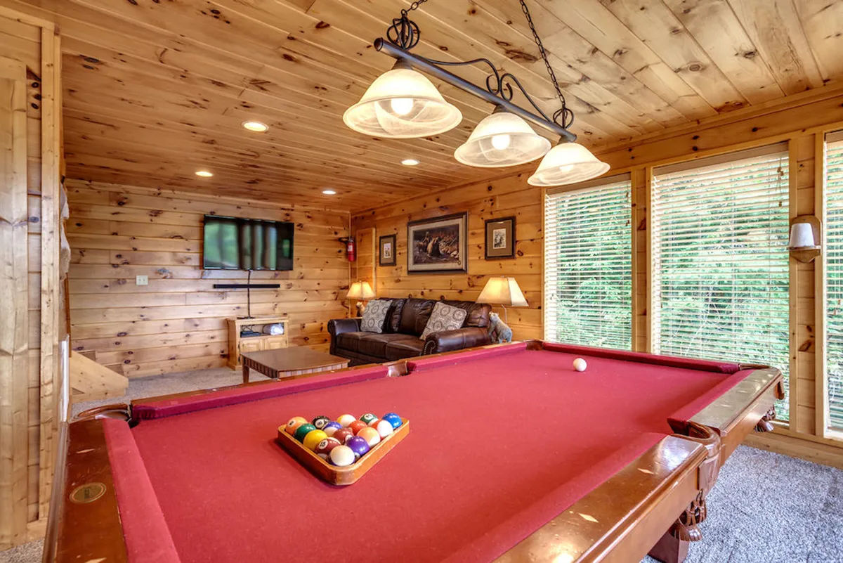 A vibrant lower-level living area featuring a pool table as the centerpiece, accompanied by a comfortable sofa and a smart TV. Large windows brighten the space, creating a fun and inviting atmosphere ideal for socializing, playing games, or enjoying a movie night.